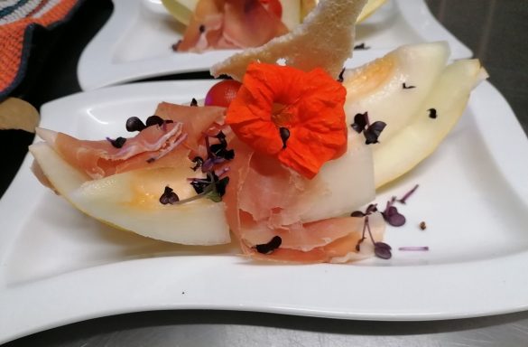 Melone mit Schinken, Blüte und essbaren Blättern auf einem weißen Teller.