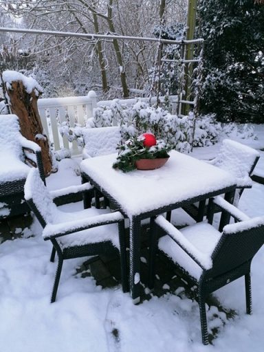 Ein mit Schnee bedeckter Tisch und Stühle im Wintergarten, dekoriert mit einem Weihnachtskranz.