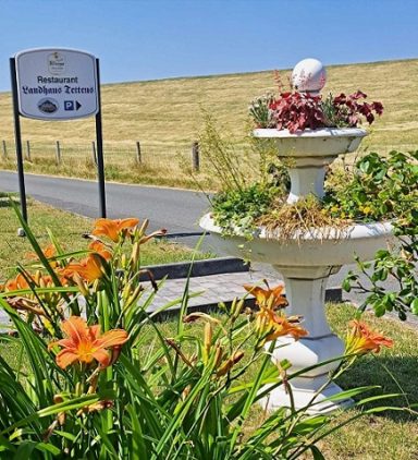 Blumenbeet mit orangefarbenen Lilien und einem Brunnen, im Hintergrund ein Schild.