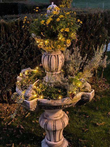 Eine dekorative Steinsäule mit gelben Blumen und Lichtern, umgeben von grünen Pflanzen.