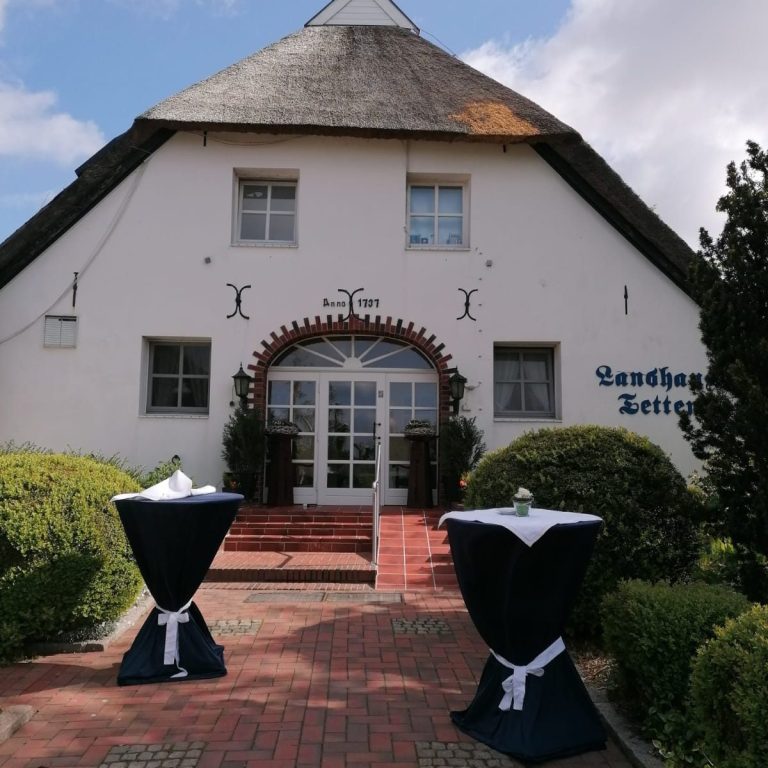 Helles Gebäude mit Reetdach und zwei gedeckten Stehtischen im Vordergrund.