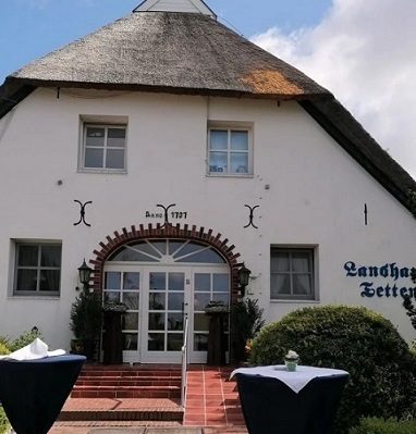Landhaus Eingang mit Reetdach und Eingangstür, umgeben von Büschen und Stehtischen.